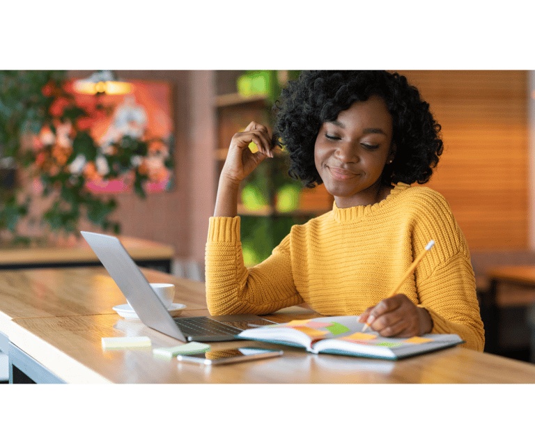 Femme souriante en pull jaune, écrivant dans un carnet avec un ordinateur