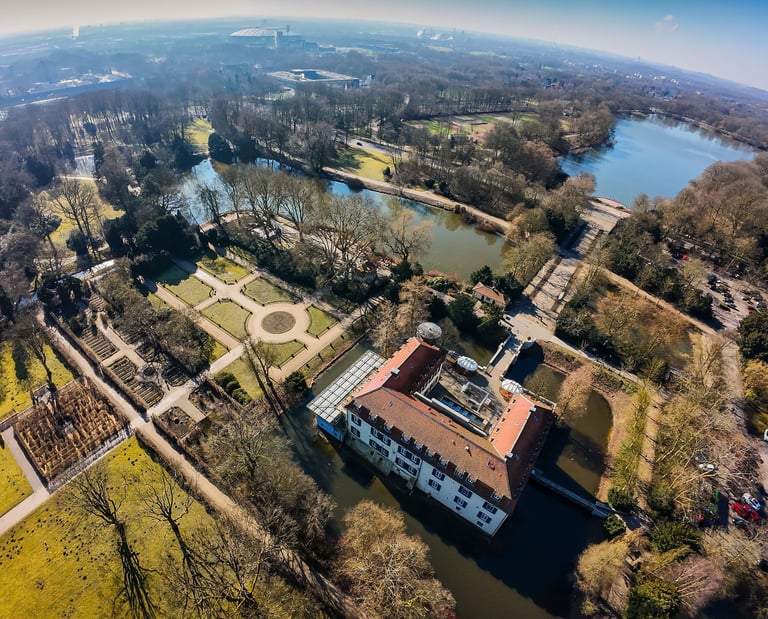 Aerial view of a historic moated castle surrounded by lush gardens, walking paths, and scenic lakes in a city park.