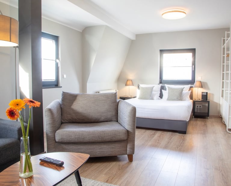 Modern hotel studio apartment with grey armchairs, king size bed, and bright hardwood floors.