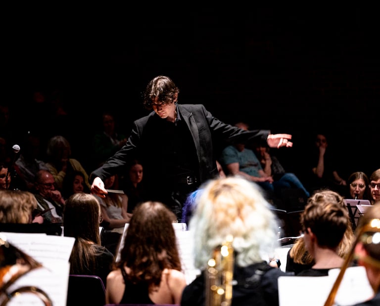 Conductor in black, with arms spread, amid heads of orchestra-members.