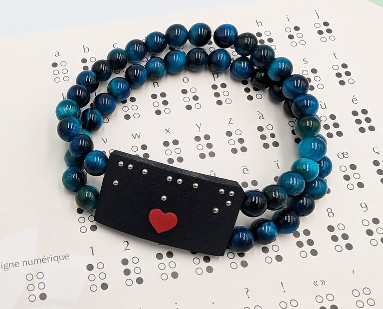 Bracelet perle bleue avec une plaque en braille et un cœur rouge, posé sur un alphabet braille.