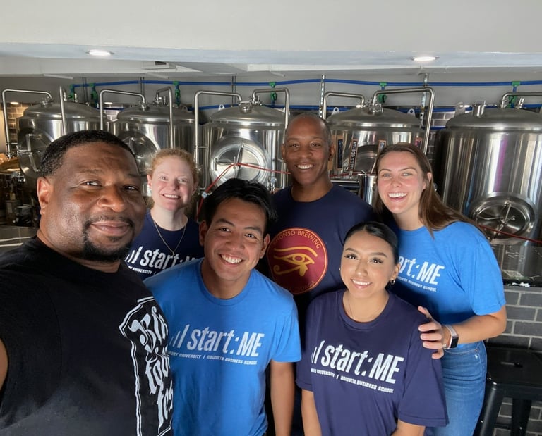 A group of diverse entrepreneurs smiling in a brewery with large stainless steel fermentation tanks.