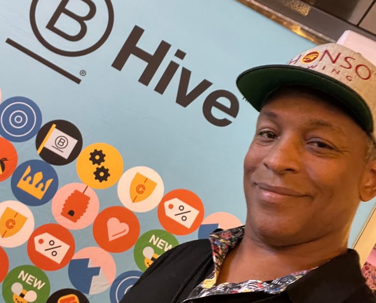 A man in a baseball cap smiling in front of a blue B Hive brand wall with colorful icons.