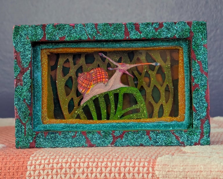 Diorama box featuring a colorful snail crawling on a leave in front of glittered layered leaf and floura affects.