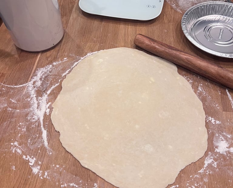 Rolled out pie crust dough on a floured wooden table with a rolling pin and baking supplies.
