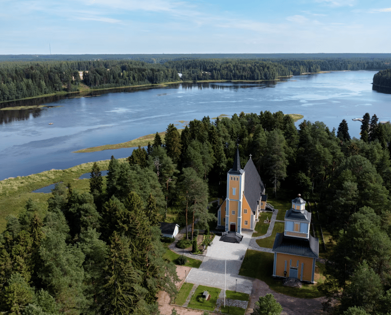 Aerial view of Muhos wooden church surrounded by forest and river, showcasing a historic landmark in