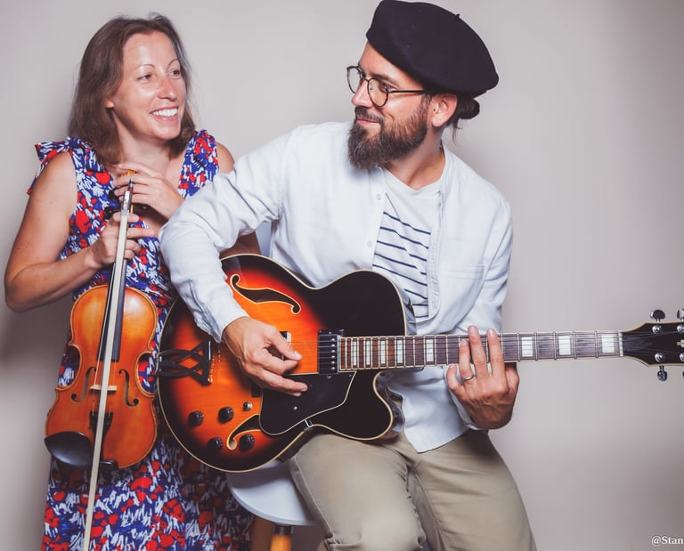 A smiling man plays a sunburst hollow body electric guitar next to a female violinist.
