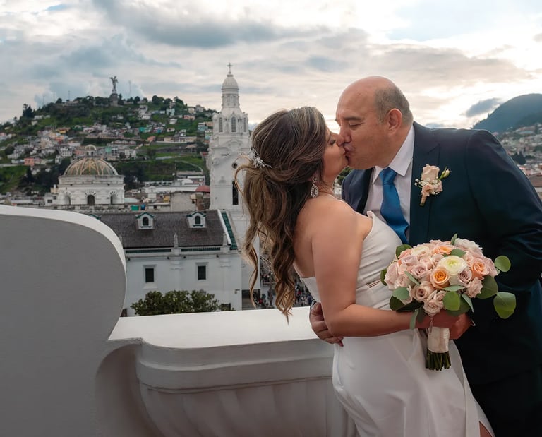 El novio besando a la novia mientras la agarra de la cintura en un terraza con vista al Panecillo en el centro histórico