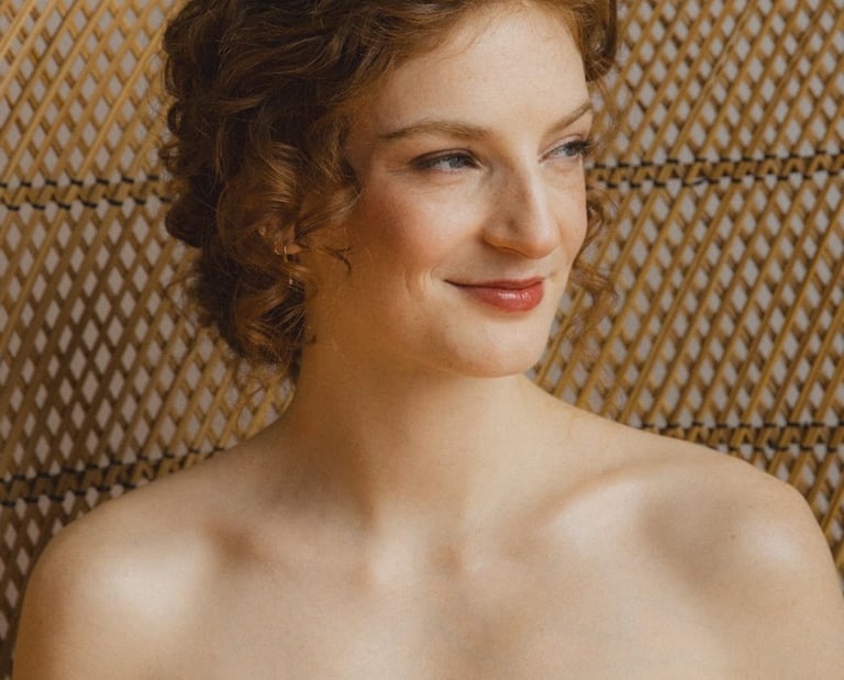 Une mariée souriante regarde vers l'extérieur. Ses cheveux sont roux et ondulés. Elle a un maquillag