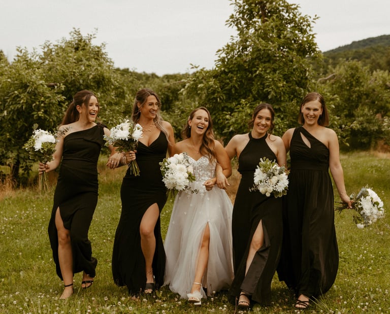 A bride in a lace gown walks and laughs with bridesmaids in black dresses at an outdoor wedding.