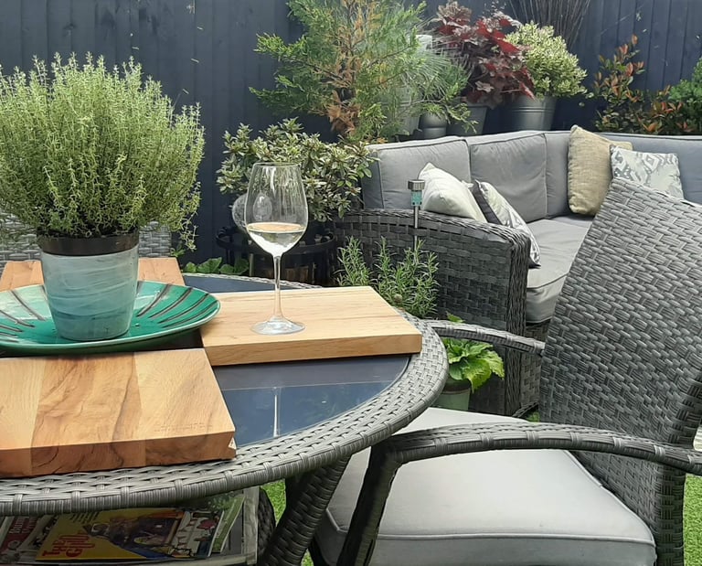 Modern garden patio with grey wicker furniture, a wine glass, and potted herbs on a glass table.