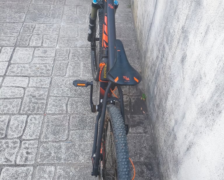 Bicicleta de montanha KTM preta e laranja vista de trás, com selim e pneus largos, apoiada junto a p
