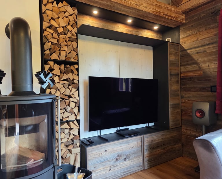 Wohnwand aus Altholz mit TV-Nische, integriertem Brennholzregal und Beleuchtung neben Kaminofen.