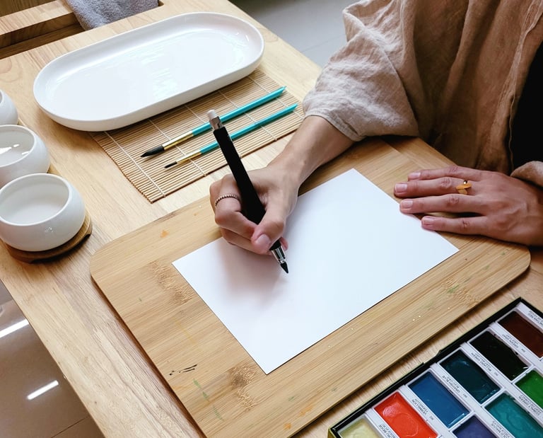 An artist drawing on white paper with a black pen, surrounded by watercolor paints and brushes.