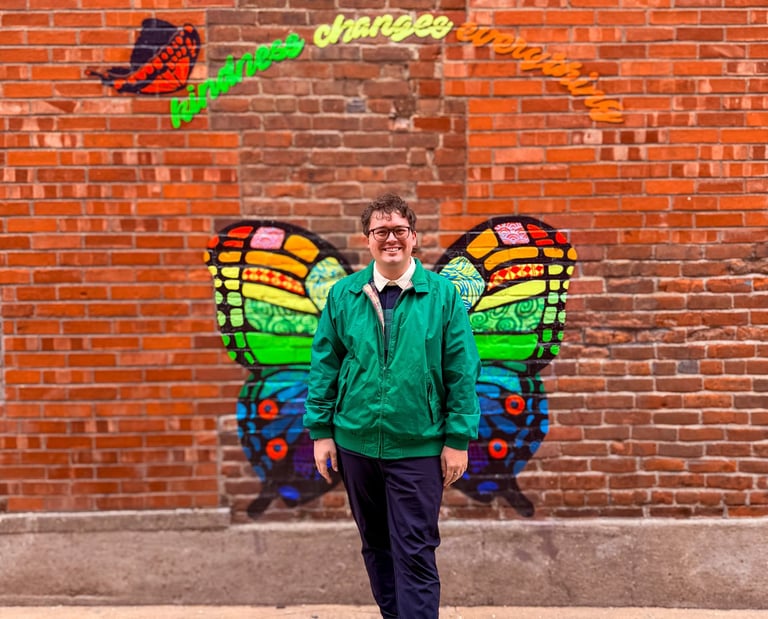 Smiling man in a green jacket posing before a colorful butterfly mural on a red brick wall.