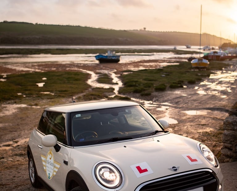 White Mini Cooper driving school car with L-plates parked at sunset by a scenic river estuary.