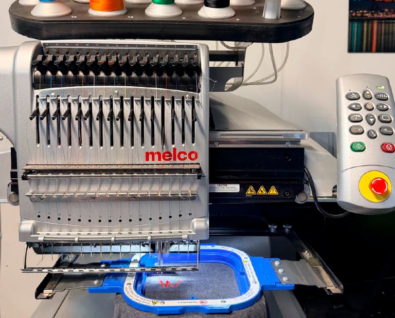Professional Melco embroidery machine stitching a red logo on grey fabric with colorful thread spools.