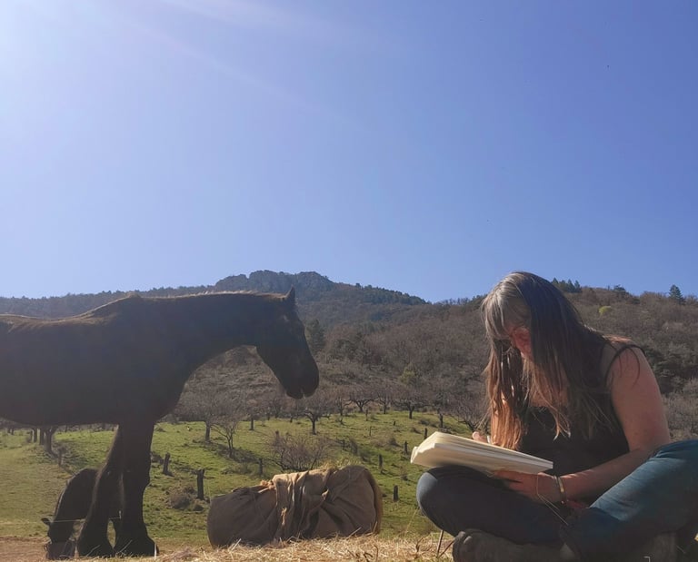 a woman sitting on the grass with a book in her hands and a horse behind her