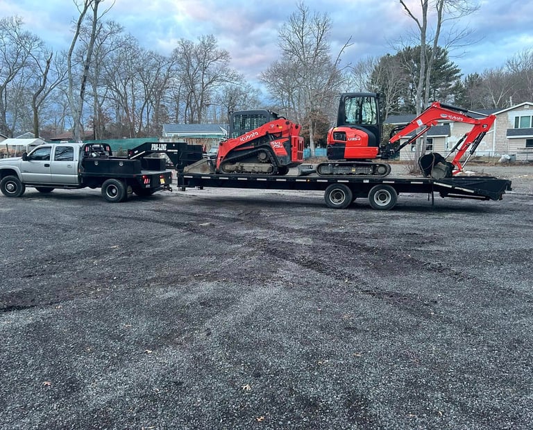 Flatbed truck towing a trailer with a Kubota skid steer and mini excavator on a gravel lot.