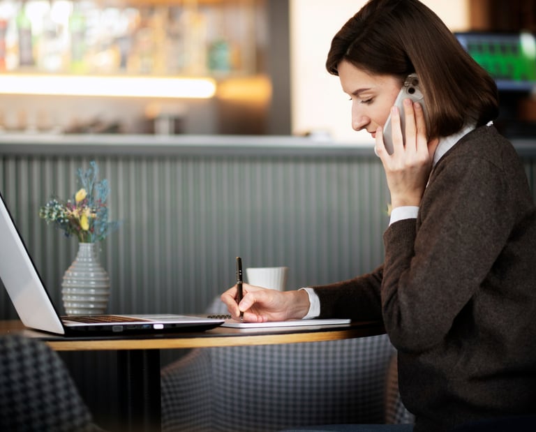 Professional woman talking on her smartphone and taking notes while working on a laptop in a cafe.