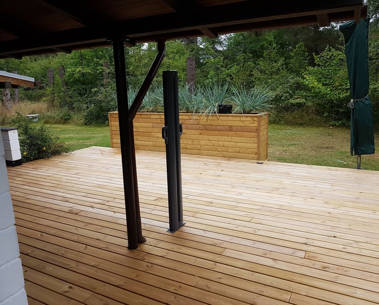 En moderne udendørs træterrasse med en stor plantekasse og en frodig grøn skov i baggrunden.