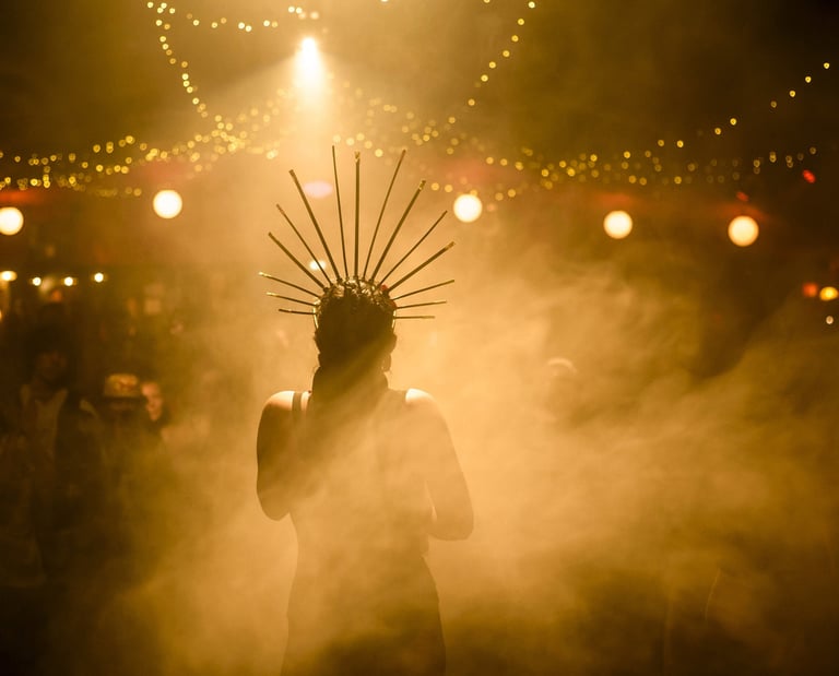A performer wearing a gold spike crown standing in a hazy club with warm stage lighting and fairy lights.