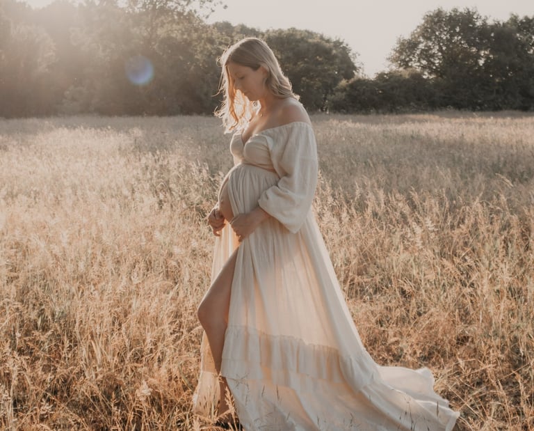 A pregnant woman in a flowing maternity dress poses in a golden sunlit meadow for an outdoor maternity photoshoot.