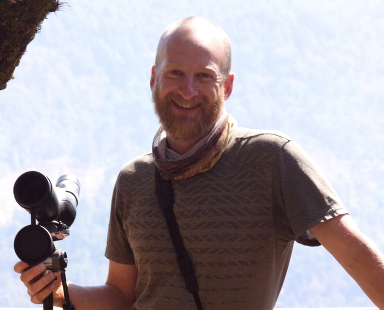 San Cristobal bird guide Benedict Simmons smiling with telescope, with binoculars in front hillside 