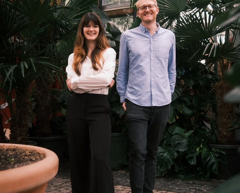 A smiling man and woman standing together in a lush indoor greenhouse surrounded by tropical plants.