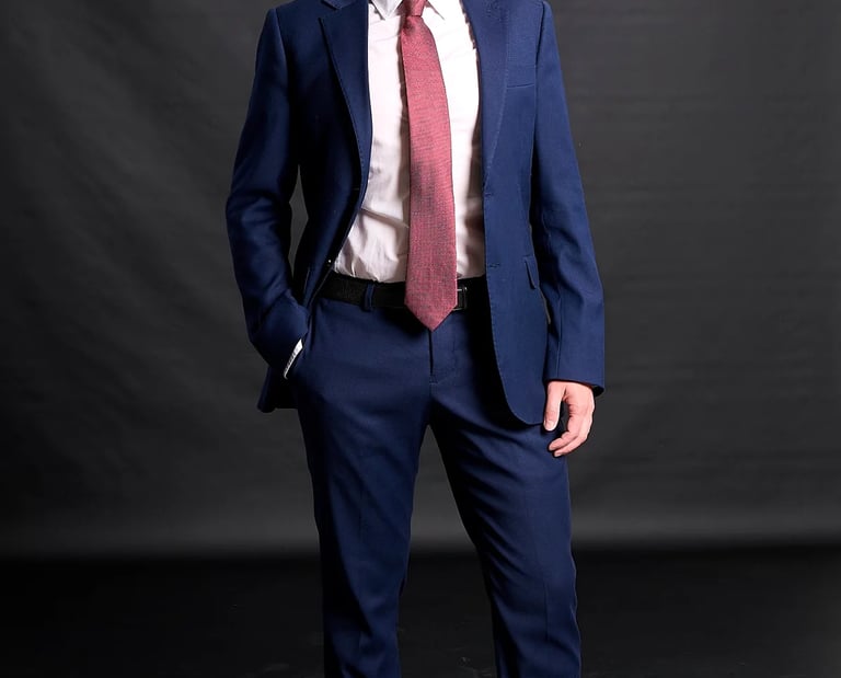 Professional portrait of a young man wearing a navy blue suit and red tie against a dark background.