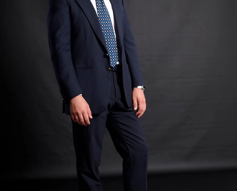 Professional man in a navy blue suit and patterned tie posing for a corporate headshot against a black background.