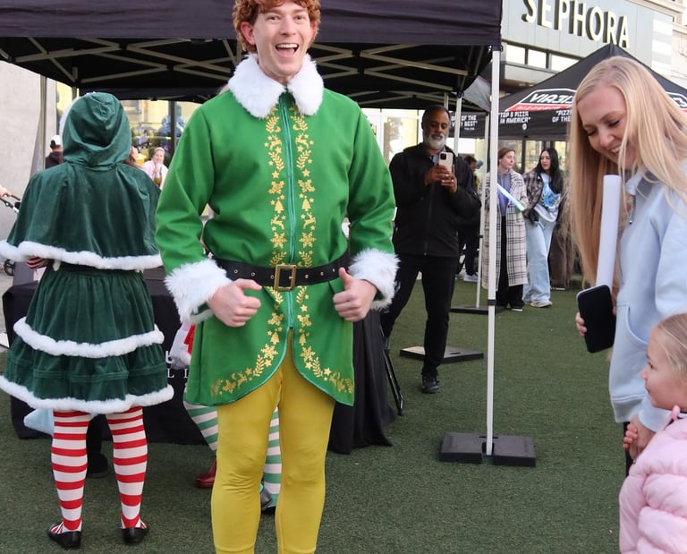 Man dressed in a Buddy the Elf costume gives a thumbs up during a festive holiday event at an outdoor mall.