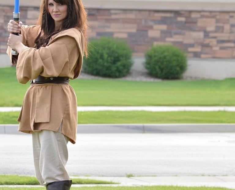 A woman in a brown Jedi cosplay costume holding a glowing blue lightsaber outdoors.