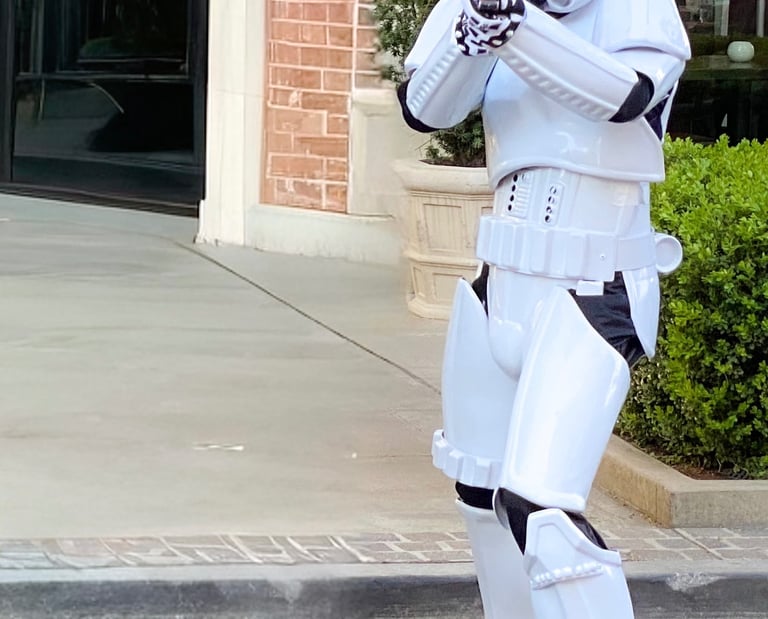 A Star Wars Stormtrooper in full white armor aims a blaster rifle while standing on a sidewalk.