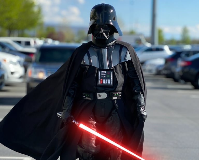A person in a realistic Darth Vader cosplay costume holding a glowing red lightsaber in a sunny parking lot.
