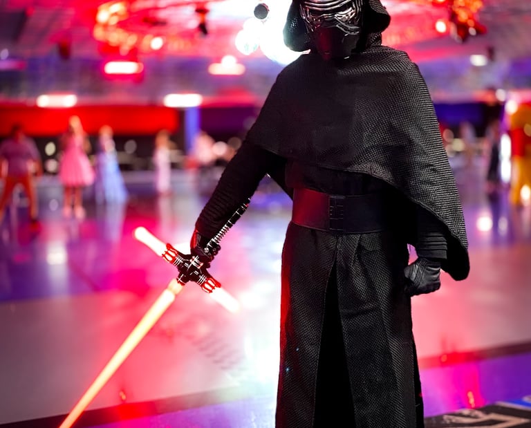 A person in a Kylo Ren cosplay costume holding a glowing red crossguard lightsaber at a roller rink.