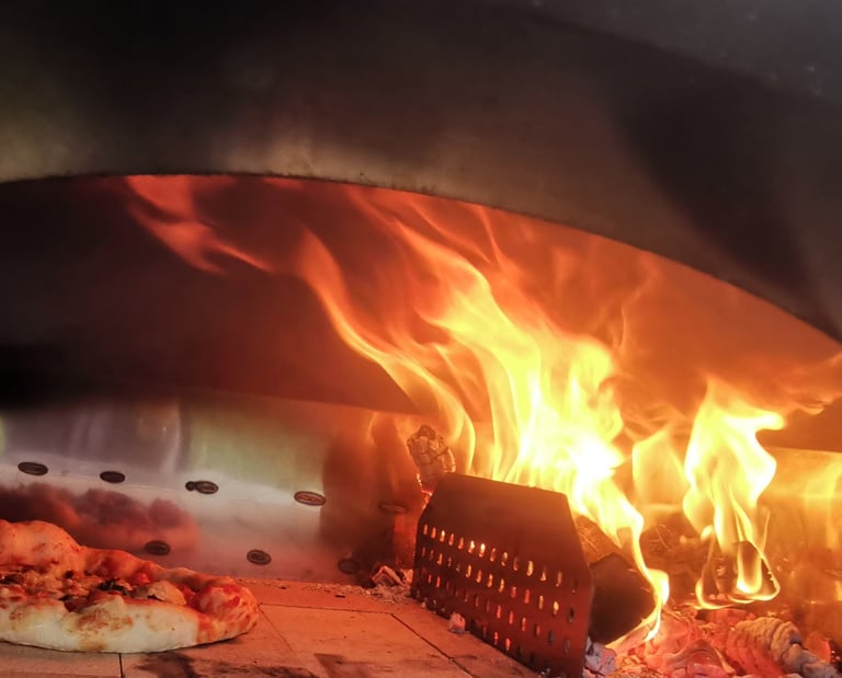 A fresh pizza baking inside a wood-fired outdoor oven with bright orange flames.