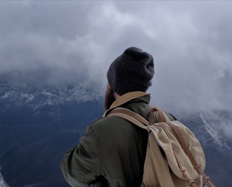A male hiker with a backpack and beanie overlooking a cloudy mountain range in winter.