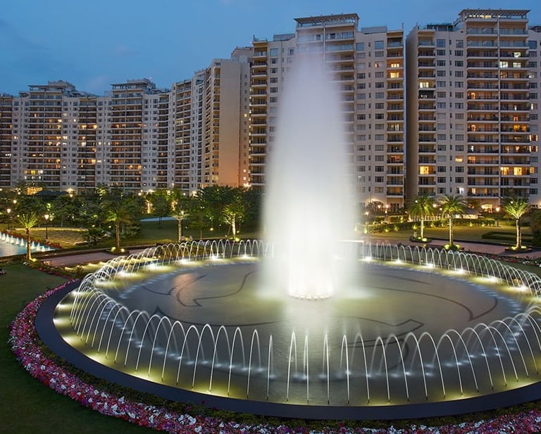 Illuminated circular water fountain in front of luxury high-rise apartment buildings at dusk.