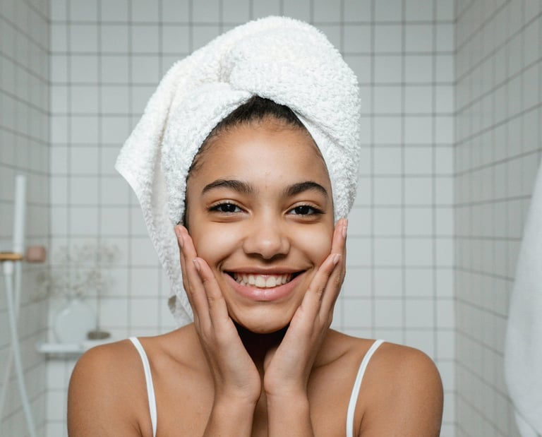 A smiling woman with a white towel head wrap shows off her glowing skin during a bathroom skincare routine.
