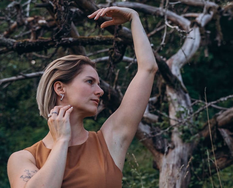 A blonde woman in a brown dress poses gracefully with raised arms in front of an old gnarled tree.