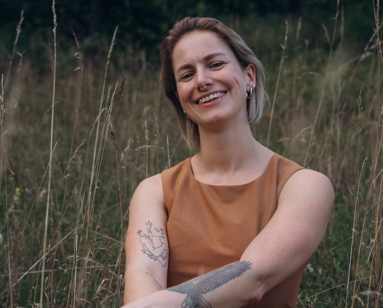 Smiling woman with tattoos wearing a brown dress sitting in a field of tall grass.