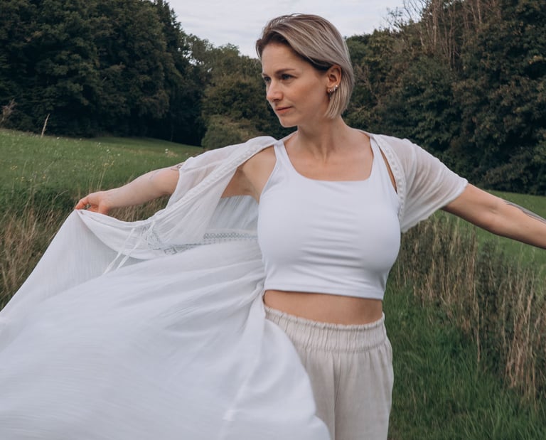 A blonde woman in white boho clothing poses in a green meadow field with trees in the background.