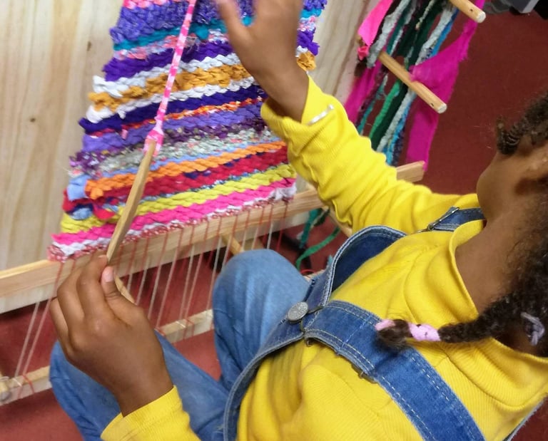 a young girl sits on a chair and weaves colorful rag rug on a weaving loom