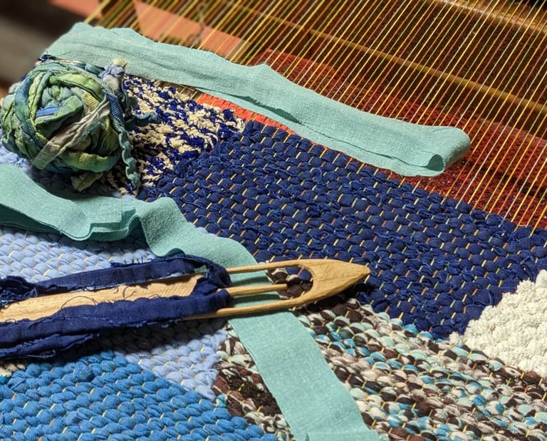 a weaving loom with a wooden shuttle, a ball of rag threads and a rag rug in shades of blue in progr