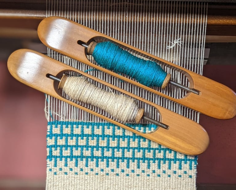 Blue and white krokbragd weaving is currently being done on a weaving loom with wooden shuttles and 
