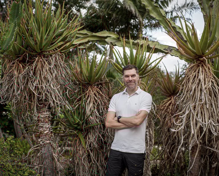 Man stands in a lush botanical garden surrounded by tall aloe and cactus plants, Madeira lifestyle and natural greenery.
