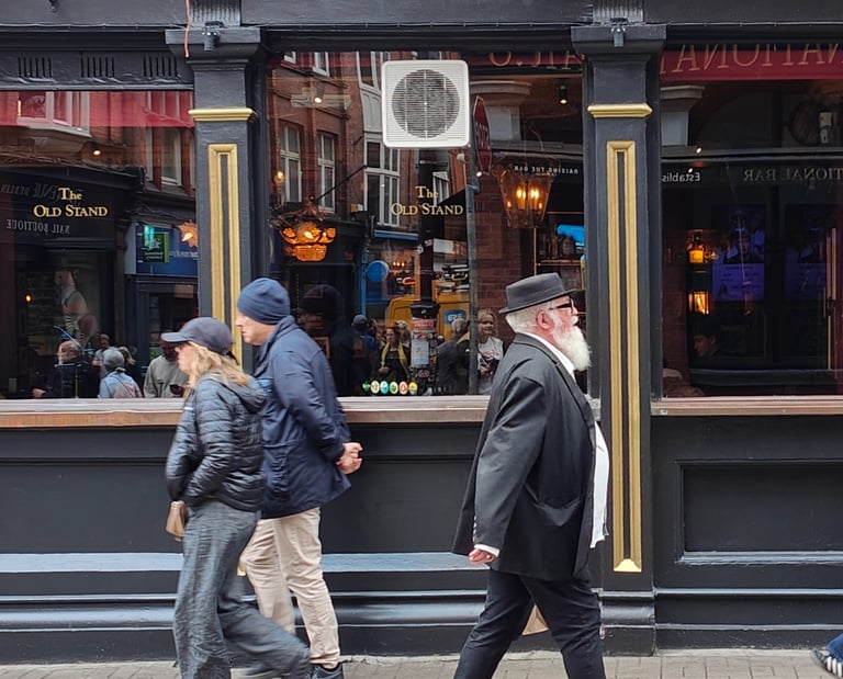 A bearded man in black hat and coat walks past The Old Stand pub; two others walk by, flowers above the sign, stone street.