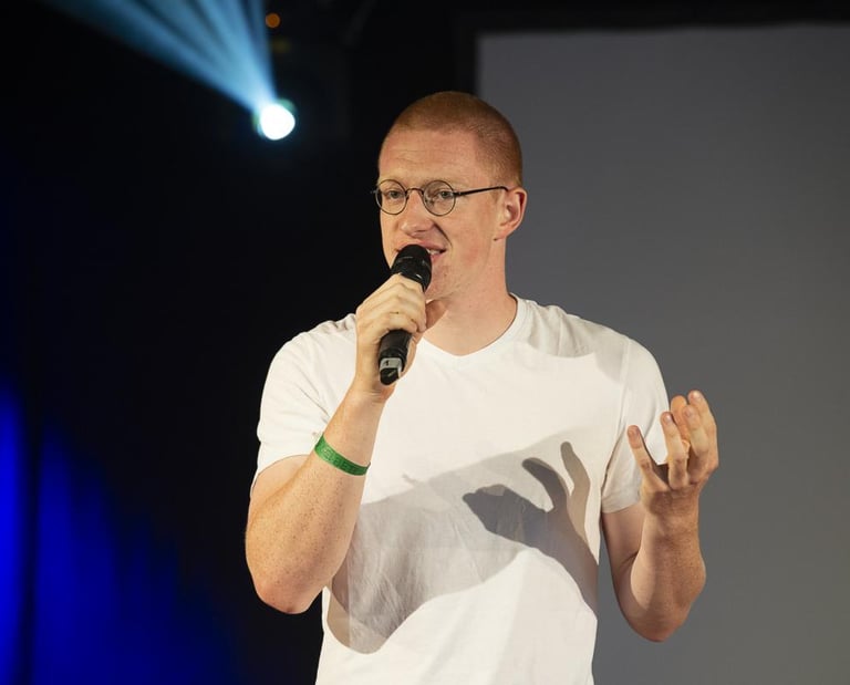 Przemysław Żuk, kurz Przemek, steht auf einer Bühne und spricht in ein Mikrofon. Er gestikuliert. Im Hintergrund Bühnenlicht.