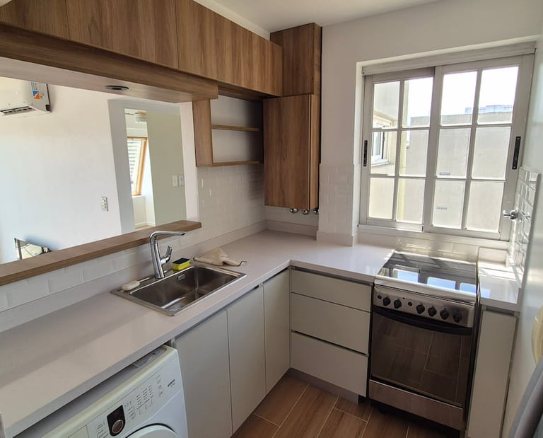 Modern small apartment kitchen with white countertops, wooden cabinets, and stainless steel appliances.